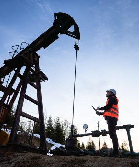 Side view snapshot of an oil engineer wearing orange vest and a helmet, looking at the oil rig and making some notes against beautiful evening sky. Concept of oil extraction and petroleum industry.